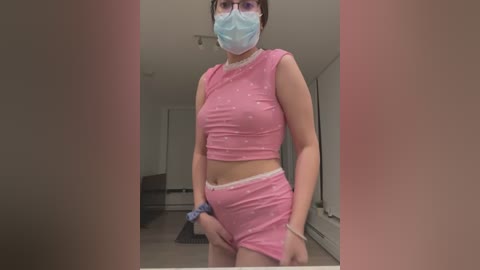 Media: Video of a woman wearing a blue face mask, pink crop top, and matching shorts, standing in a modern, minimalist room.