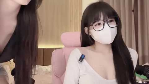 Media: Video of an East Asian woman with long black hair, wearing glasses, white mask, and a white top, sitting on a pink chair in a room with beige curtains and a bed.