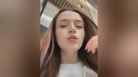 Media: Video of a young Caucasian woman with fair skin, long brown hair, and light makeup. She wears a white top, slightly parted lips, and looks directly at the camera. Background shows a modern indoor setting with wooden slats.