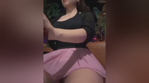 Media: A video captures a young woman with light skin, wearing a black top and pink pleated skirt, seated in a dimly lit room with greenery and red lights in the background.