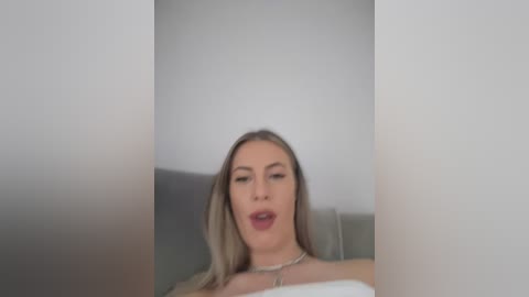 A video of a light-skinned woman with long blonde hair, wearing a silver necklace, and a white top, with a neutral expression, standing in a minimalistic room with white walls.