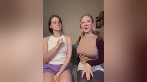 Media: Video of two young Caucasian women with fair skin, sitting side-by-side, wearing casual attire: one in a white tank top and purple shorts, the other in a brown long-sleeve and grey shorts. They smile, with the background showing a beige wall and a wooden shelf.