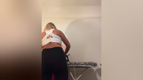 Media: A video of a blonde woman in a white sports bra and black leggings ironing clothes on a white ironing board in a dimly lit room.