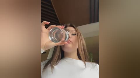 Media: A video of an Asian woman with straight, shoulder-length brown hair, wearing a white shirt, drinking from a clear glass water bottle, set against a modern interior with brown wooden beams and a white ceiling.