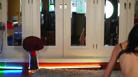 Media: Video of a woman with long black hair, partially nude, sitting on a shaggy rug in a modern living room. Behind her, white double doors reflect colorful LED lights, and a potted plant sits on the floor.