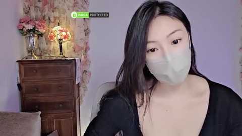 Media: Video of an East Asian woman with long black hair, wearing a face mask, black top, and seated indoors. Background features a wooden dresser, floral wallpaper, and a lit lamp.