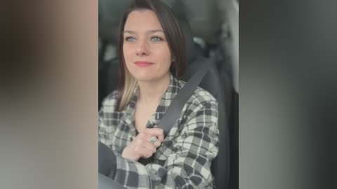 Video of a fair-skinned woman with long brown hair, wearing a black and white plaid shirt, buckled in a car seat, with blurred background.