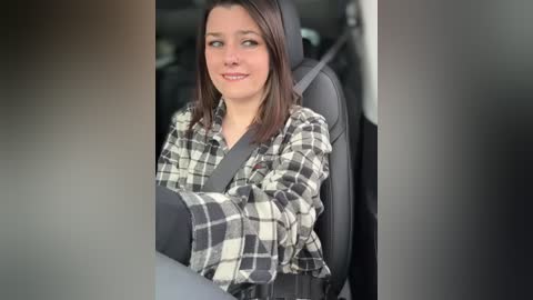 Media: Video of a young woman with straight brown hair, wearing a gray plaid flannel shirt and black leggings, seated in a car with a gray seatbelt.