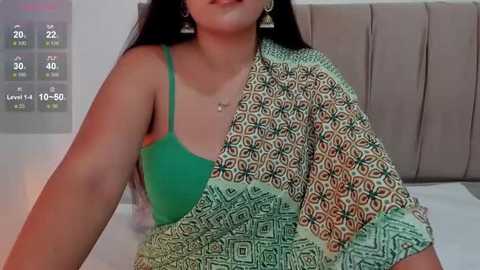 Media: Video of a South Asian woman with long black hair, wearing a green bra and a patterned, multi-colored saree, sitting on a bed with a beige headboard.