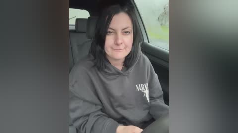 Media: Video of a woman with straight black hair, wearing a dark gray hoodie with white text, sitting in a car. The background shows a blurred view of a bicycle and green grass.