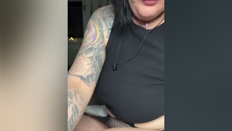 Media: Video of a woman with tattoos on her arm, wearing a black crop top and fishnet stockings. Her face is partially visible, with lips painted in dark red lipstick.