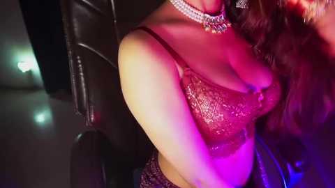 Media: Video of a woman in a black leather jacket, wearing a glittering, sheer, pink top, with a necklace and earrings, sitting in a dimly lit room with purple and green lighting.