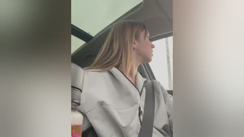 Media: Video of a Caucasian woman with straight, shoulder-length blonde hair, wearing a light grey jacket, seated in a car with a seatbelt, looking out the window. The background shows a blurry, beige interior.