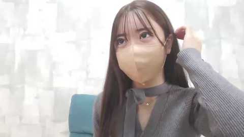 Media: A video of an Asian woman with long, straight brown hair, wearing a grey ribbed sweater and a beige mask, adjusting her hair in a modern, minimalist office setting with white patterned wallpaper.