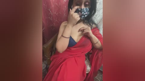 Media: Video of a South Asian woman with medium skin tone, black hair, and medium build. She wears a red saree, revealing a blue bra, and a black mask. Background features a red curtain and floral wallpaper.