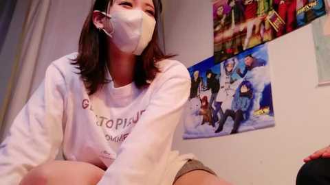 Media: Video of an Asian woman with long black hair, wearing a white \"GOTOPLESS\" sweatshirt, face mask, and headphones, sitting on a bed. Background features colorful posters and a plush toy.