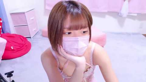 Media: Video of an Asian woman with short brown hair, wearing a pink mask, pink lace lingerie, and a red bra, sitting on a bed in a pastel-colored room.