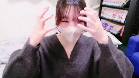 Media: Video of a young woman with short, dark hair, wearing a gray sweater, a face mask, and black-framed glasses, adjusting her glasses. Background includes a blue chair, white wall, and bookshelves.