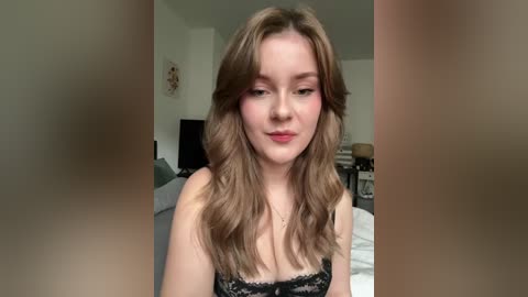 Video of a fair-skinned, young woman with long, wavy brown hair, wearing a black lace bralette. Background shows a cluttered bedroom with a bed, desk, and wall art.