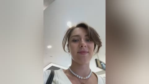 Media: Video of a smiling, light-skinned woman with short brown hair, wearing a white blouse and pearl necklace, standing in a modern, well-lit room with white walls and recessed lighting.