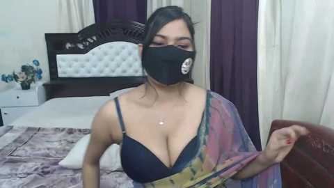 Media: Video of a South Asian woman with medium skin tone, dark hair, wearing a black face mask, blue bra, and multicolored shawl, standing in a bedroom with purple curtains, white walls, and a dark wooden headboard.