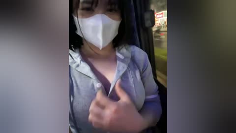 Media: A video of an Asian woman with medium skin tone, wearing a white face mask, gray top, and dark hair, giving a thumbs-up gesture. Background shows blurred car interior with a visible sign.