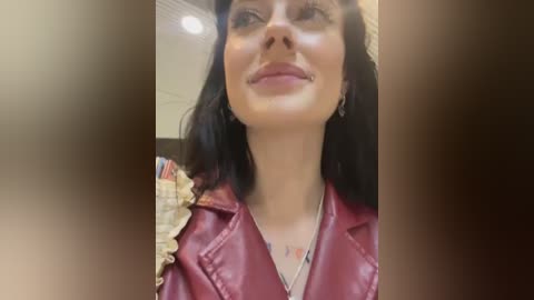 Media: Video of a smiling woman with long black hair, wearing a red leather jacket, standing in a dimly lit room with beige walls and a bookshelf in the background.