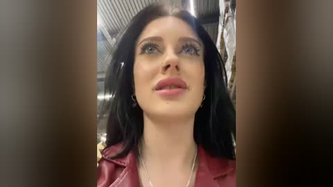 Media: Video of a fair-skinned woman with long black hair, wearing a red leather jacket and bold makeup, in an industrial setting with metal beams and hanging clothes in the background.