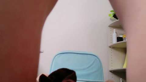 Media: A video of a woman in a light blue chair, seen from a low angle, with a potted plant and shelf in the background.