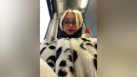 Media: A video of a blonde woman with glasses and a black-and-white cow print jacket, standing in a bus, wearing a neutral expression.