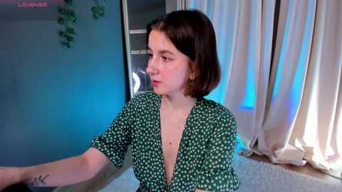 Media: A video of a young woman with a short bob haircut, fair skin, and green polka-dot dress, sitting indoors with blue and white curtains, a window, and a plant in the background.