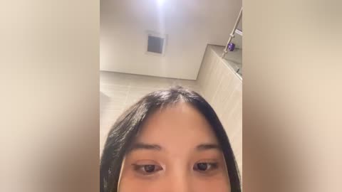 Media: A close-up video of a young woman with long, straight black hair, light skin, and large brown eyes, taken from a low angle. The background features a modern bathroom with beige tiles, a showerhead, and a vent.