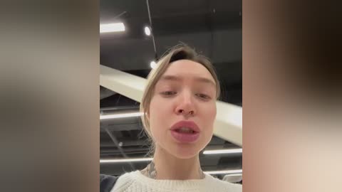 Media: A video of a young woman with fair skin and blonde hair, wearing a white sweater, sticking out her tongue in an exaggerated manner. The background shows a modern, industrial interior with dark walls and a white staircase.