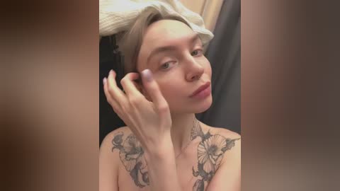 Media: Video of a young, fair-skinned woman with platinum blonde hair wrapped in a towel, applying makeup with a hand adorned with a detailed floral tattoo on her collarbone.