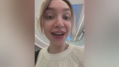 Media: Video of a young woman with fair skin and blonde hair, smiling with a nose ring, wearing a cream knit sweater. Background shows a modern, white interior with blue accents.