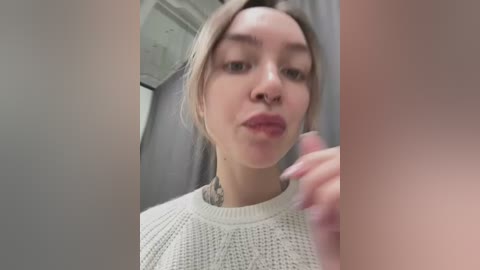 Media: Video of a young woman with light skin, light blonde hair, and a nose ring, wearing a white knit sweater. She blows a kiss and holds a toothbrush in her mouth, with a tattoo visible on her neck.