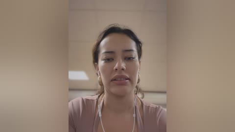 A video of a young woman with medium brown skin and dark hair in a ponytail, wearing a light pink blouse, standing in a beige-tiled corridor.