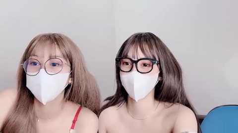 Media: Video of two East Asian women with long hair, wearing white masks and black-rimmed glasses, sitting side by side, indoors against a plain white background.