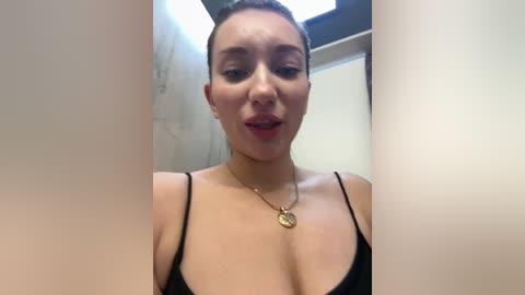 Media: Video of a light-skinned woman with short dark hair, wearing a black spaghetti strap top, standing in a dimly lit bathroom with beige walls and a window.