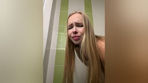 Video of a blonde woman with long, straight hair, wearing a white tank top, leaning over, looking distressed, in a bathroom with green and white striped tiles.