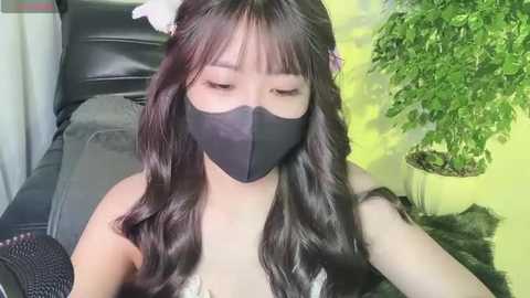 Media: Video of an East Asian woman with long, wavy black hair and fair skin, wearing a black mask and a black sleeveless top, sitting on a black leather chair, with a potted plant in the background.