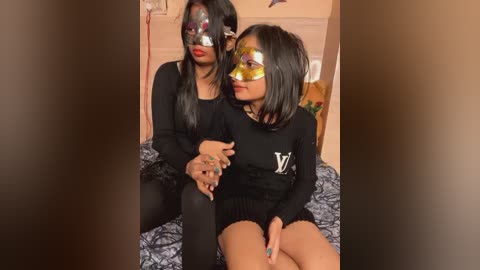 Media: Video of two women in black masks, black dresses, and short hair, sitting on a bed with floral sheets, in a warmly lit room.