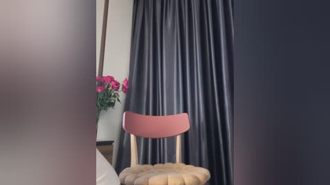 Media: Video of a minimalist bedroom with a wooden chair covered in a plush, beige cushion, set against a backdrop of dark, pleated curtains. A vase of vibrant pink flowers sits on a small table to the left.