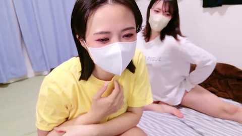 Media: Video of two Asian women in surgical masks, sitting on a bed in a hospital room. One wears a yellow shirt, the other a white one, both with medium builds.