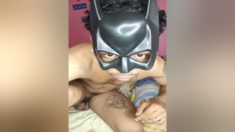 Media: A video of a nude man wearing a Batman mask, with visible tattoos and erect penis, in a dimly lit room with a pink wall and blue sheets.