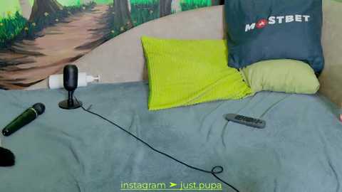 Media: Video of a bed with a green comforter, yellow pillow, and a dark pillow with \"Mostbet\" logo. A microphone, remote control, and cable are on the bed. Background features a painted forest scene. Text reads: \"Instagram Just dubs.\