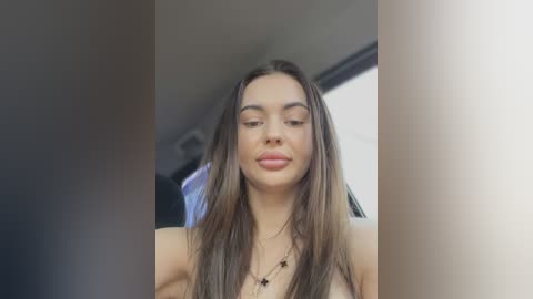 Media: Video of a young woman with long brown hair, light skin, and a closed-mouth smile, wearing a black necklace. Background features a blurred indoor setting with soft lighting.