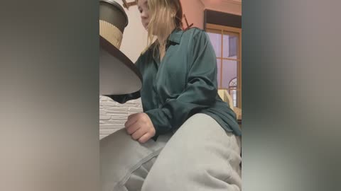 Media: Video of a blonde woman in a teal shirt and gray sweatpants, crouching under a bed with a beige headboard, in a dimly lit room with a window and a wicker basket.