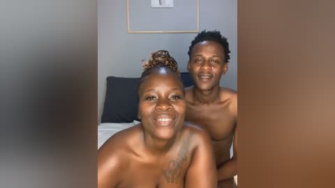 Media: Video of a smiling Black couple, both topless, in a bedroom. The woman has a curly bun, and the man has short, curly hair. Background includes a bed with a dark pillow and framed art on the wall.