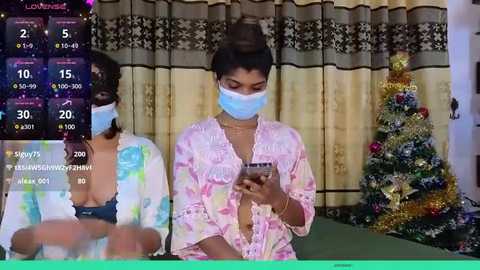 Media: Video of a young woman in a pink floral robe, wearing a blue surgical mask, looking at her phone. Background includes a Christmas tree and curtains.